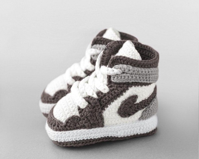 K&ouml;nnte beinhalten: Ein Paar handgefertigte Baby-H&auml;kel-Sneaker im High-Top-Stil. Die Schuhe haben eine wei&szlig;e Basis mit braunen und grauen Akzenten, einschlie&szlig;lich des ikonischen Swoosh-Designs. Wei&szlig;e Schn&uuml;rsenkel vervollst&auml;ndigen den Look. Ein einzigartiges Geschenk.