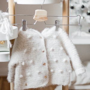 Pode incluir: Um casaco de tric&ocirc; branco para beb&ecirc; com fecho de bot&atilde;o e pequenos pompons brancos por toda a superf&iacute;cie. O casaco est&aacute; pendurado em um rack de madeira com um cabide de madeira.