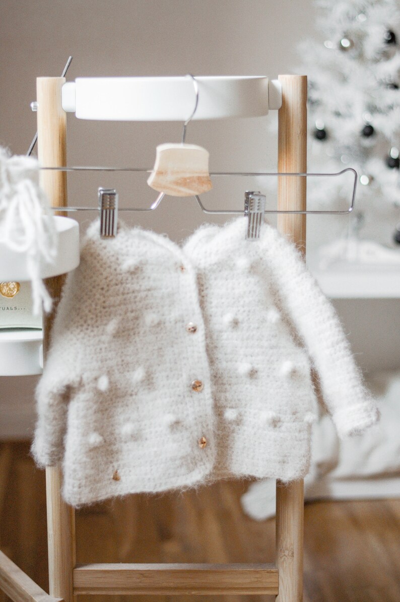 Pode incluir: Um casaco de tric&ocirc; branco para beb&ecirc; com fecho de bot&atilde;o e pequenos pompons brancos por toda a superf&iacute;cie. O casaco est&aacute; pendurado em um rack de madeira com um cabide de madeira.