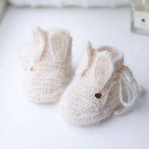 Pode incluir: Um par de sapatinhos de beb&ecirc; de tric&ocirc; cor creme com orelhas de coelho e detalhes de cora&ccedil;&atilde;o dourado.