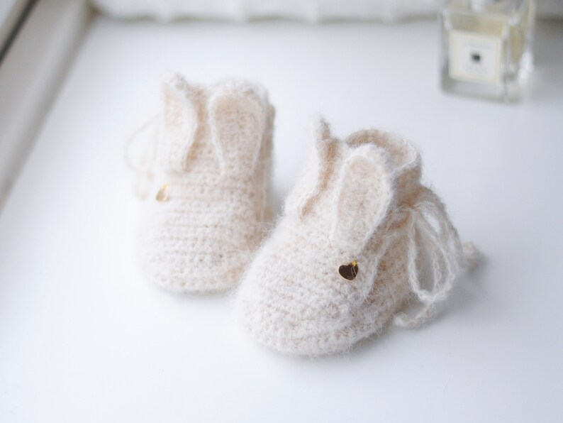 Pode incluir: Um par de sapatinhos de beb&ecirc; de tric&ocirc; cor creme com orelhas de coelho e detalhes de cora&ccedil;&atilde;o dourado.