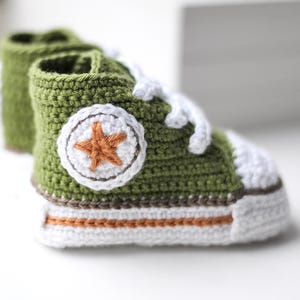以下が含まれることがあります： 緑色の毛糸で編まれたベビーシューズ。白い紐と、側面に茶色の星が付いています。