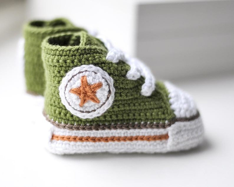 以下が含まれることがあります： 緑色の毛糸で編まれたベビーシューズ。白い紐と、側面に茶色の星が付いています。