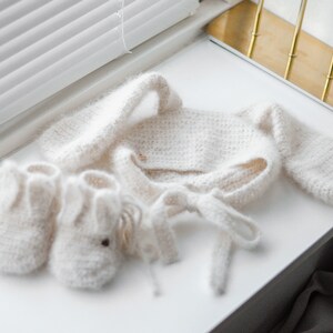 Pode incluir: Um conjunto de beb&eacute; de malha branca com um par de botinhas a condizer. O conjunto inclui um cardigan de manga comprida com um la&ccedil;o na cintura e um chap&eacute;u a condizer.