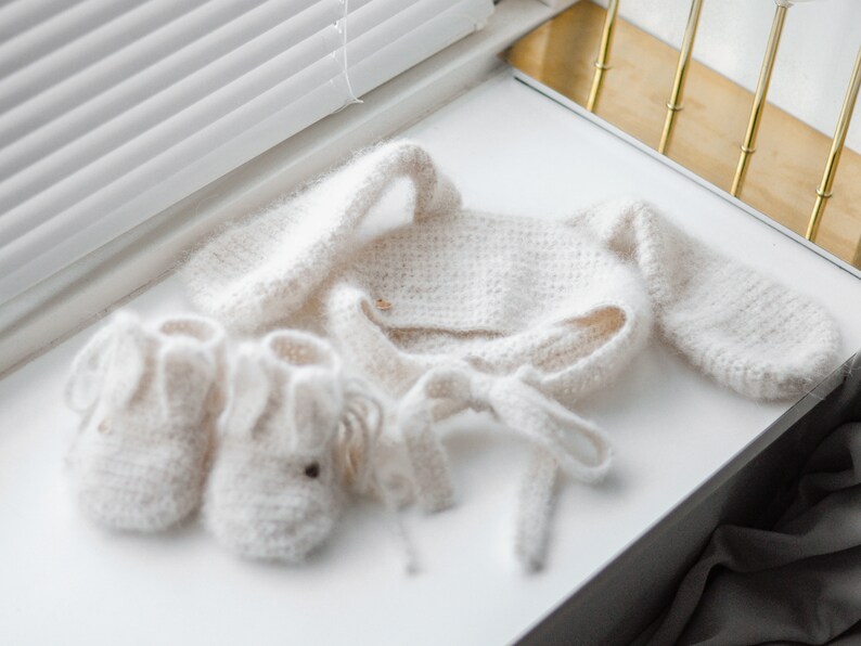 Pode incluir: Um conjunto de beb&eacute; de malha branca com um par de botinhas a condizer. O conjunto inclui um cardigan de manga comprida com um la&ccedil;o na cintura e um chap&eacute;u a condizer.