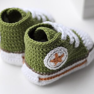 以下が含まれることがあります： 緑色の毛糸で編まれたベビーシューズのペア。白い紐と、側面に茶色の星が付いています。