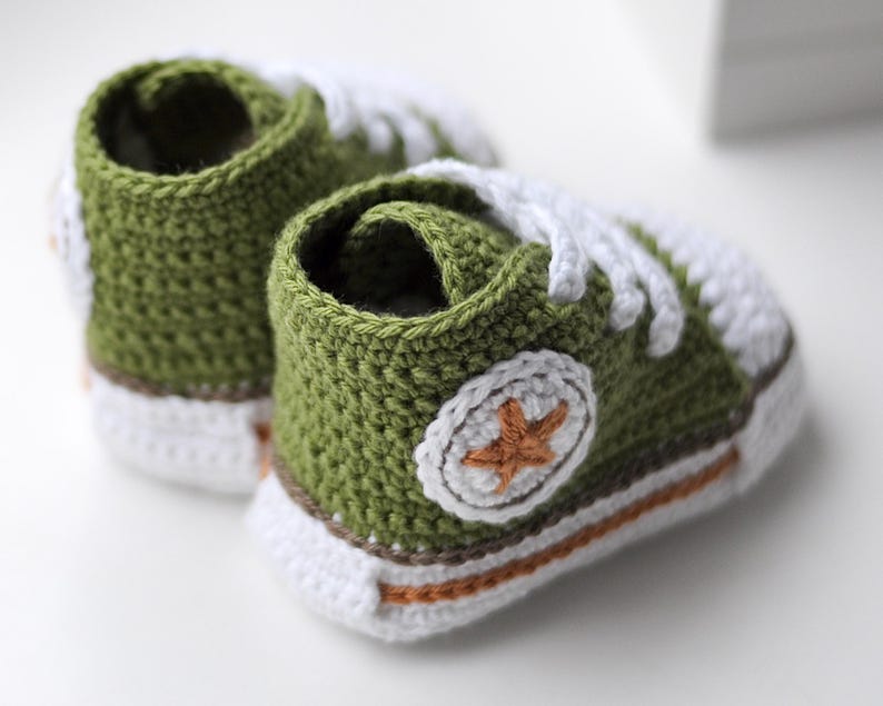 以下が含まれることがあります： 緑色の毛糸で編まれたベビーシューズのペア。白い紐と、側面に茶色の星が付いています。