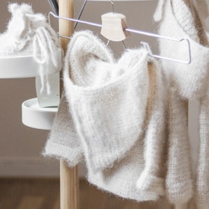 Pode incluir: Tr&ecirc;s pe&ccedil;as de vestu&aacute;rio de croch&ecirc; branco penduradas em um suporte de madeira. As pe&ccedil;as s&atilde;o um su&eacute;ter com capuz, uma echarpe e um par de botinhas.