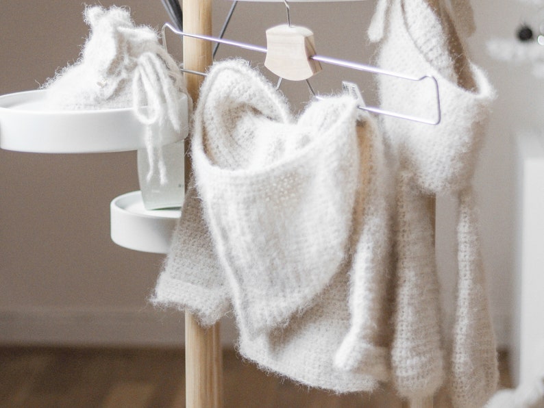 Pode incluir: Tr&ecirc;s pe&ccedil;as de vestu&aacute;rio de croch&ecirc; branco penduradas em um suporte de madeira. As pe&ccedil;as s&atilde;o um su&eacute;ter com capuz, uma echarpe e um par de botinhas.