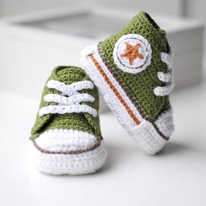 以下が含まれることがあります： 緑色の毛糸で編まれたベビーシューズのペア。白い紐と、前面に茶色の星が付いている。