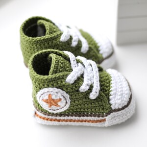 以下が含まれることがあります： 緑色の編みぐるみベビーシューズのペア。白いレースとオレンジ色の星が側面に付いています。靴の底は白とオレンジの縁取りがされています。