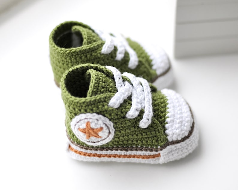 以下が含まれることがあります： 緑色の編みぐるみベビーシューズのペア。白いレースとオレンジ色の星が側面に付いています。靴の底は白とオレンジの縁取りがされています。