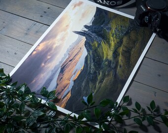 May include: A panoramic photograph of a rocky mountain landscape with a dramatic sky. The mountain is covered in green vegetation and has a sharp peak. The sky is a mix of blue, gray, and orange clouds.