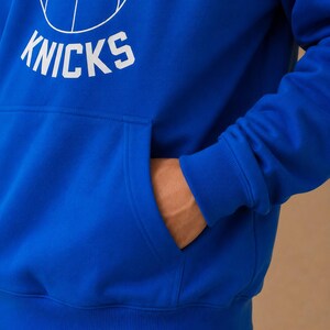 May include: Royal blue hoodie with a white basketball graphic and the word "KNICKS". The hoodie features a front pocket and a drawstring hood. The NBA logo is visible on the basketball graphic.