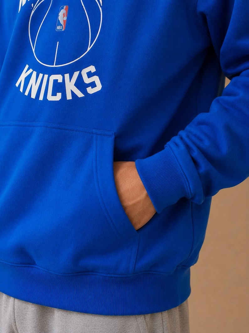 May include: Royal blue hoodie with a white basketball graphic and the word "KNICKS". The hoodie features a front pocket and a drawstring hood. The NBA logo is visible on the basketball graphic.