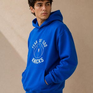 May include: Royal blue hoodie with a front pocket and a hood. The hoodie features the text "FEAR OF GOD" above a basketball graphic and the word "KNICKS" below. The person is wearing gray sweatpants.