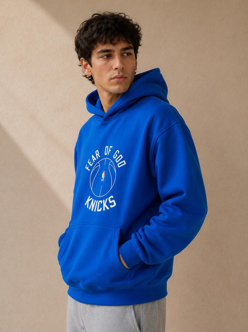 May include: Royal blue hoodie with a front pocket and a hood. The hoodie features the text "FEAR OF GOD" above a basketball graphic and the word "KNICKS" below. The person is wearing gray sweatpants.