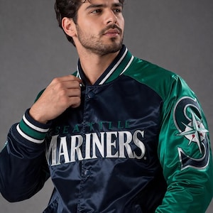 May include: A navy blue and green satin Seattle Mariners baseball jacket. The jacket features the team name in white lettering across the chest and a team logo on the sleeve. The collar and cuffs have white and green stripes.