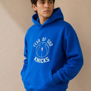May include: Royal blue hoodie with a front kangaroo pocket and a drawstring hood. The hoodie features a white graphic with the text "FEAR OF GOD" above a basketball and the word "KNICKS" below.
