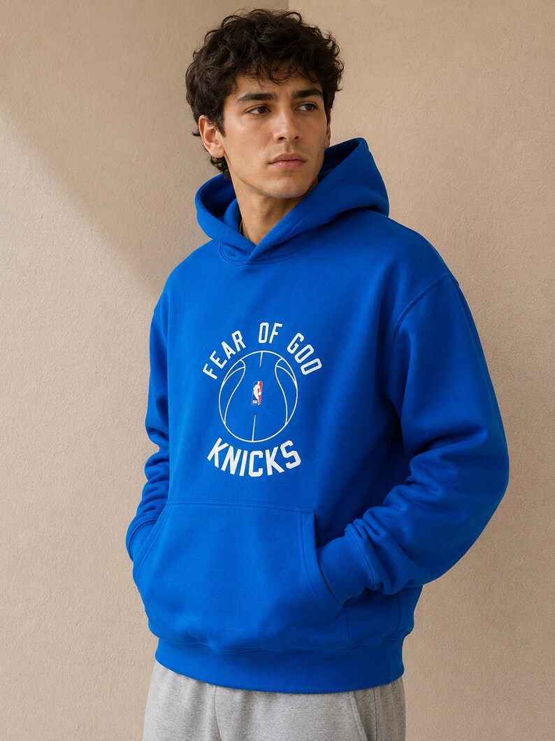 May include: Royal blue hoodie with a front kangaroo pocket and a drawstring hood. The hoodie features a white graphic with the text "FEAR OF GOD" above a basketball and the word "KNICKS" below.