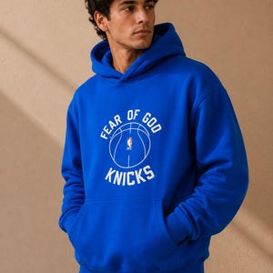 May include: Royal blue hoodie with a front kangaroo pocket. The hoodie features the text "FEAR OF GOD KNICKS" in white, along with a basketball graphic. The hoodie is made of a soft material and has a hood with drawstrings.
