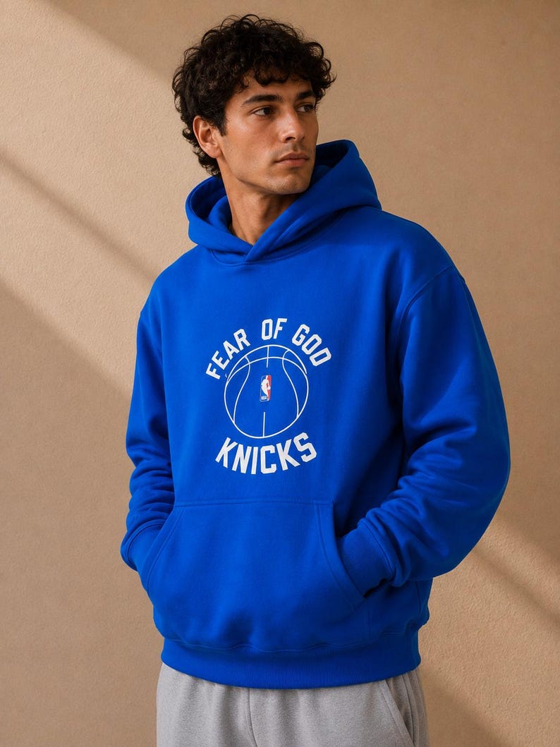 May include: Royal blue hoodie with a front kangaroo pocket. The hoodie features the text "FEAR OF GOD KNICKS" in white, along with a basketball graphic. The hoodie is made of a soft material and has a hood with drawstrings.