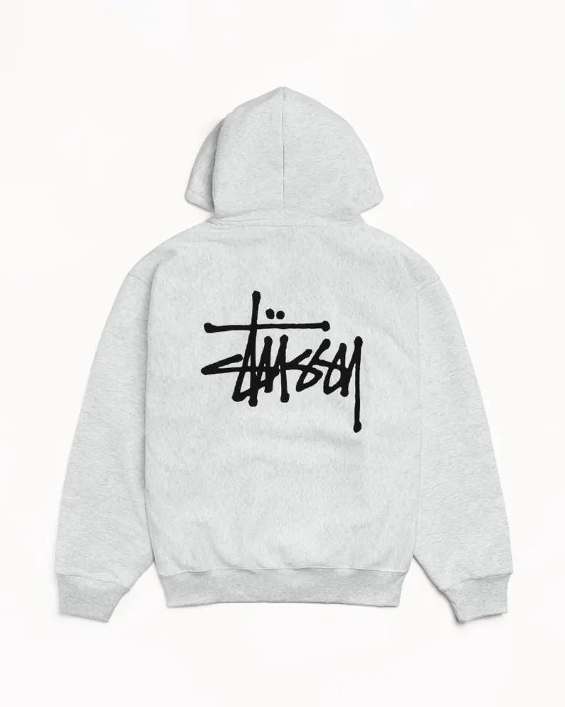 May include: A light gray hoodie with a drawstring hood and long sleeves. The back of the hoodie features a black logo. The hoodie is made of a soft, comfortable material, perfect for casual wear.