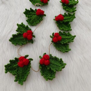 May include: A festive crocheted garland with green holly leaves and vibrant red berries. The leaves are intricately stitched, and the berries are grouped together. The garland is strung on natural twine, ideal for Christmas decorations.