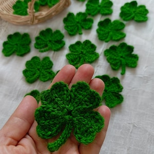May include: A hand holding a green crocheted four-leaf clover. The clover is made with a soft, textured yarn and has a stem.