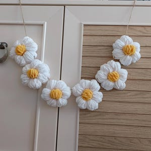 May include: A garland of six white crocheted daisies with yellow centers hangs on a white door with wood paneling.