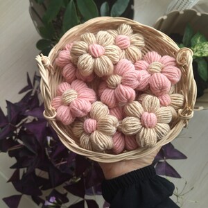May include: A wicker basket filled with handmade crocheted flowers in shades of pink and beige. The flowers are arranged in a circular pattern and have a delicate, feminine look.