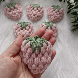 Könnte beinhalten: Fünf rosa gehäkelte Erdbeeren mit grünen gehäkelten Blättern. Die Erdbeeren sind in einem Halbkreis angeordnet, wobei sich eine Erdbeere in der Mitte der Hand befindet.
