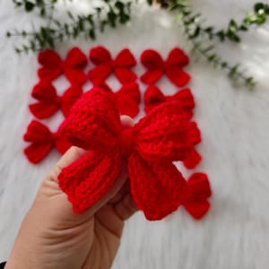 Pode incluir: Um close-up de um laço de tricô vermelho. O laço é feito à mão e tem uma textura macia e fofa. É perfeito para adicionar um toque de alegria festiva a qualquer projeto.