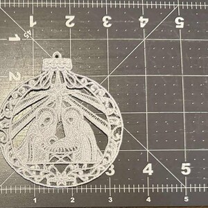 以下が含まれることがあります： ボール型のグレーのレーザーカットクリスマスオーナメント。装飾品は、装飾的なフレーム内の詳細な降誕シーンを特徴としています。デザインには、マリア、ヨセフ、幼いイエスの姿が含まれています。装飾品は薄く繊細な素材で作られています。