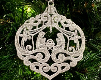 Puede incluir: Adorno navideño de encaje blanco con una escena de la natividad. El adorno tiene forma de lágrima y tiene detalles intrincados.