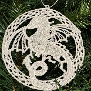 May include: A white, circular dragon ornament with intricate lace detailing. The dragon is centered within a decorative frame, featuring outstretched wings and a curled tail. The ornament hangs from a silver string, set against a backdrop of green pine needles.