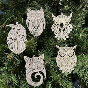 May include: Five white lace owl ornaments hanging on a green Christmas tree. Each owl has a unique design, with intricate details and varying wing and body shapes. The ornaments are made of a delicate, open-weave material.