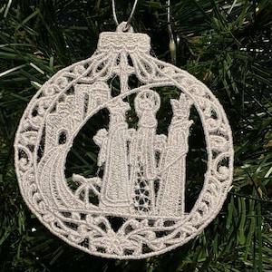 May include: A white, intricately designed Christmas ornament. The ornament features a detailed depiction of the Three Wise Men, crafted with delicate lace-like patterns. The ornament is suspended against a backdrop of green Christmas tree branches.