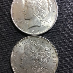 May include: Two silver Peace Dollar coins, one from 1921 and the other from 1923. Both coins feature a profile of a woman with flowing hair and the inscription "In God We Trust".