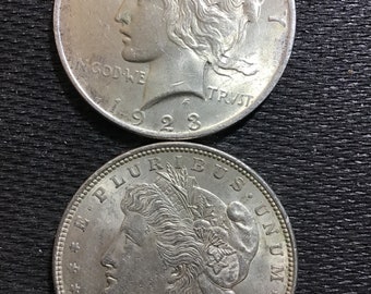 Op de afbeelding: Twee zilveren Peace Dollar-munten, een uit 1921 en de andere uit 1923. Beide munten tonen een profiel van een vrouw met golvend haar en de inscriptie "In God We Trust".