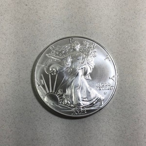 May include: A silver American Eagle coin with a depiction of Lady Liberty walking forward, holding a torch and olive branch. The coin has the inscription "In God We Trust" and the year "2023".