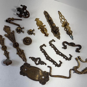Puede incluir: Una colección de herrajes para muebles de latón variados que incluye tiradores de cajón, manijas, escudos y pomos. Las piezas presentan diversos diseños ornamentados, grabados y lisos en tonos metálicos dorados y bronce.
