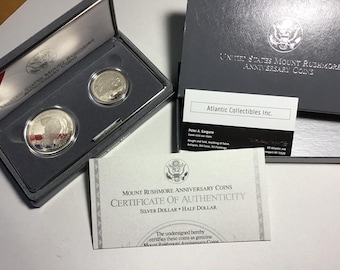 May include: A certificate of authenticity for two silver Mount Rushmore Anniversary Coins, a silver dollar and a half dollar. The certificate is in a white frame with black text. The coins are in a black velvet box with a white interior.