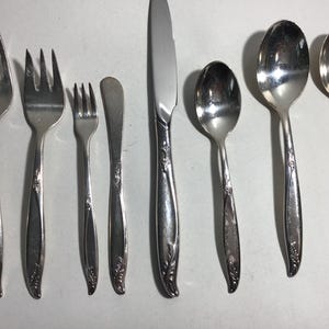 May include: A collection of silver-colored flatware, including forks, spoons, and a knife. The set features a floral design on the handles. The arrangement includes a variety of utensils for different courses, suitable for a formal dining setting.