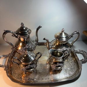 Puede incluir: Un juego de té plateado que consta de una tetera grande, una tetera más pequeña, un azucarero con tapa y una jarra para crema, todos con detalles florales en relieve, dispuestos sobre una bandeja de servir rectangular a juego.