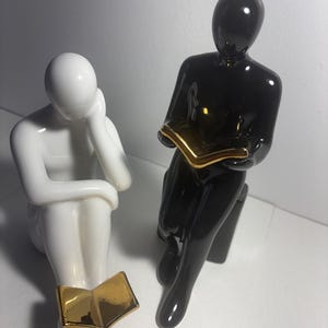 Puede incluir: Dos figuras abstractas de cerámica, una blanca brillante y otra negra brillante, ambas en posición sentada. La figura blanca apoya la cabeza en una mano, mientras que la negra sostiene un libro abierto. Ambas figuras tienen detalles dorados.