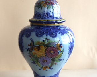 May include: A blue and white ceramic vase with a lid, decorated with floral designs in shades of yellow, red, and purple. The vase has a bulbous body and a narrow neck.