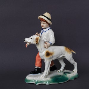 Puede incluir: Una figurita de porcelana de un niño con un sombrero de paja y pantalones rojos, sosteniendo un perro blanco y marrón con la lengua larga.