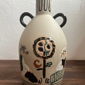 Op de afbeelding: Een beige keramische vaas met zwarte handvatten en een volkskunstontwerp met twee figuren en een boom. De vaas heeft een golvend zwart ontwerp rond de hals.