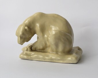 May include: A ceramic sculpture of a polar bear sitting on a block of ice. The bear is light beige in color and has a realistic appearance.